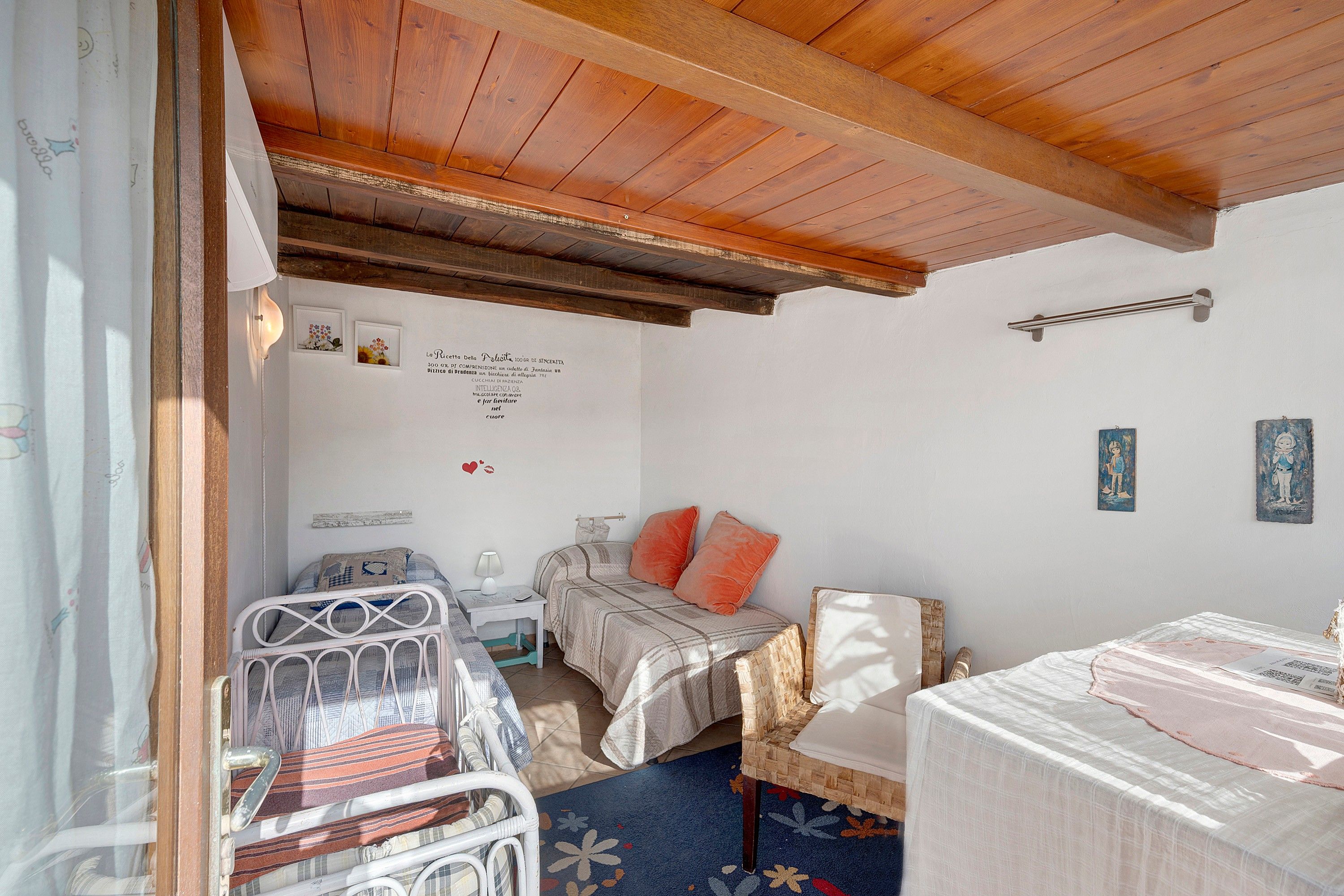 A room with a wooden ceiling, a bed with a patterned bedspread, a small table with a lamp, and a chair.