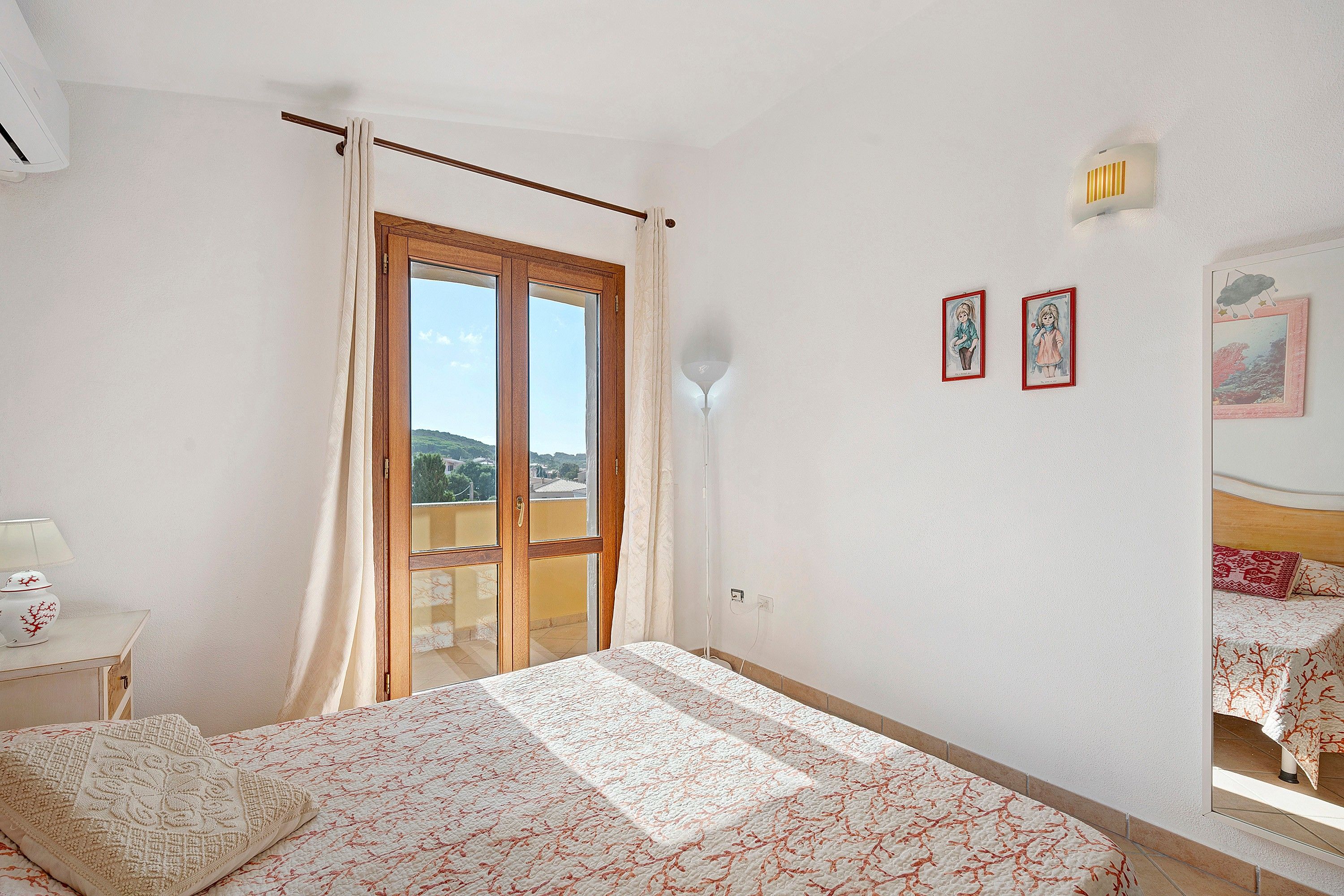 Ein Schlafzimmer mit einem Bett, einem Nachttisch, einer Lampe, einem Fenster mit Vorhängen und zwei gerahmten Bildern an der Wand.