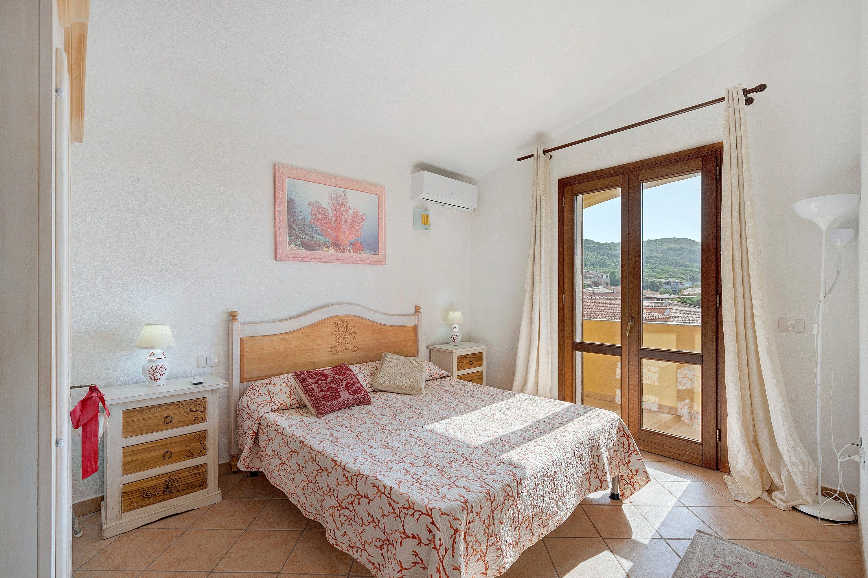Ein Schlafzimmer mit einem Bett, Nachttischen, einer Fensterbank, einer Wand-Klimaanlage, einem Fenster mit Vorhängen und Blick ins Freie.