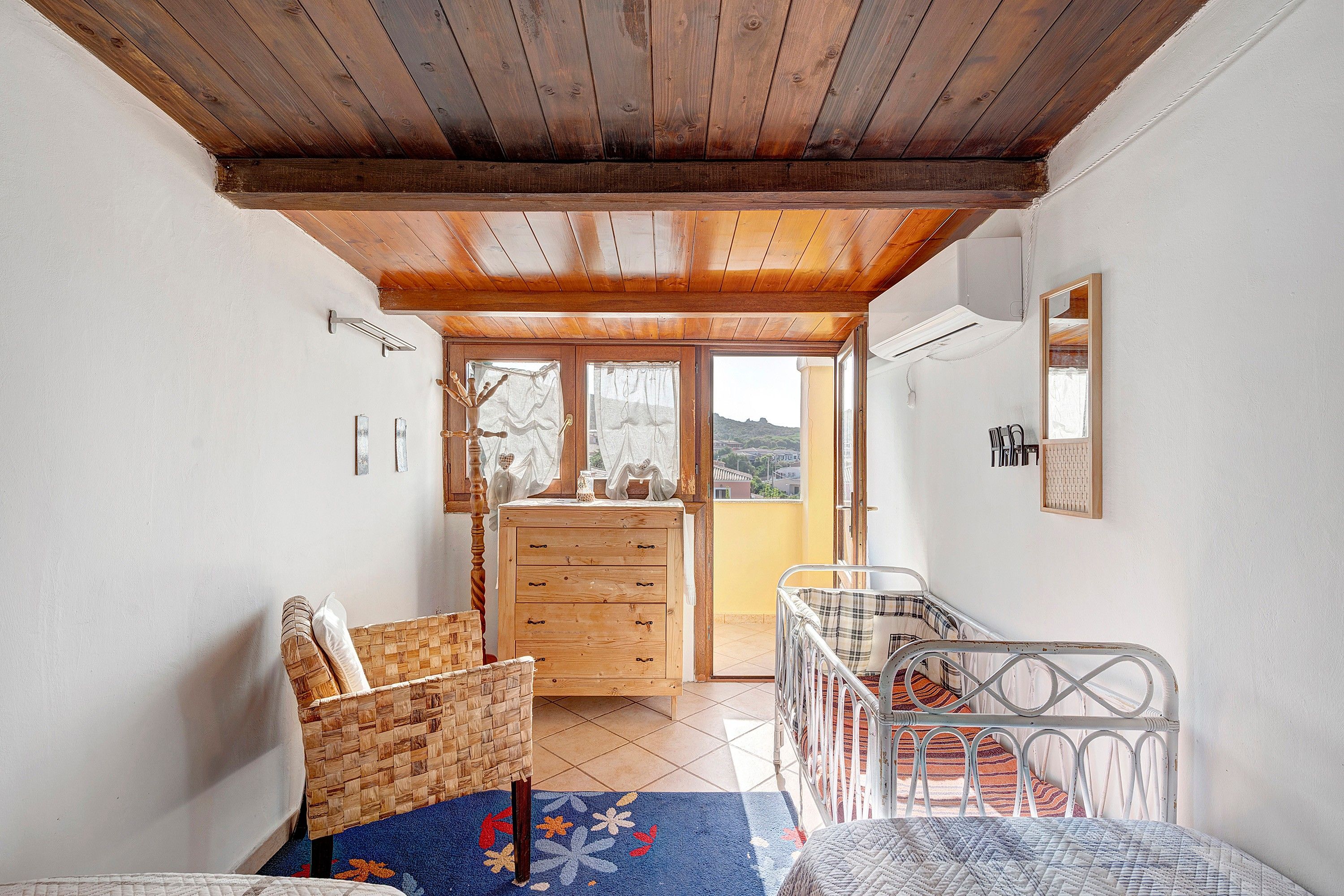 A room with a wooden ceiling, a bed with a metal frame, a wicker chair, a wooden dresser, and a view of the outside.