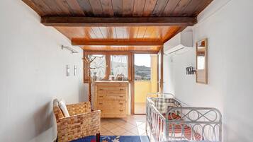 A room with a wooden ceiling, a bed with a metal frame, a wicker chair, a wooden dresser, and a view of the outside.