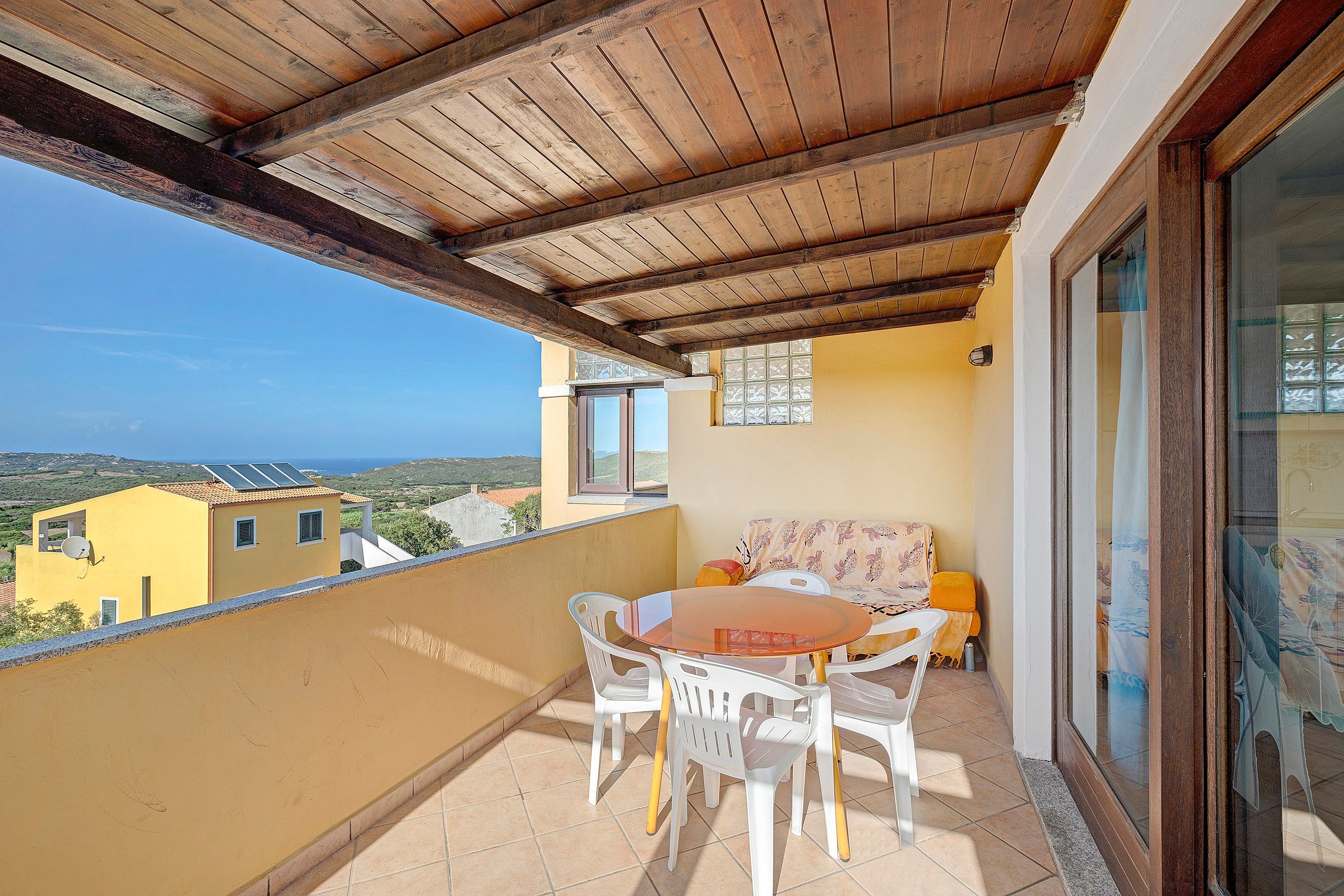 A balcony with a wooden ceiling, a table with chairs, a sofa, and a view of the sea and buildings.