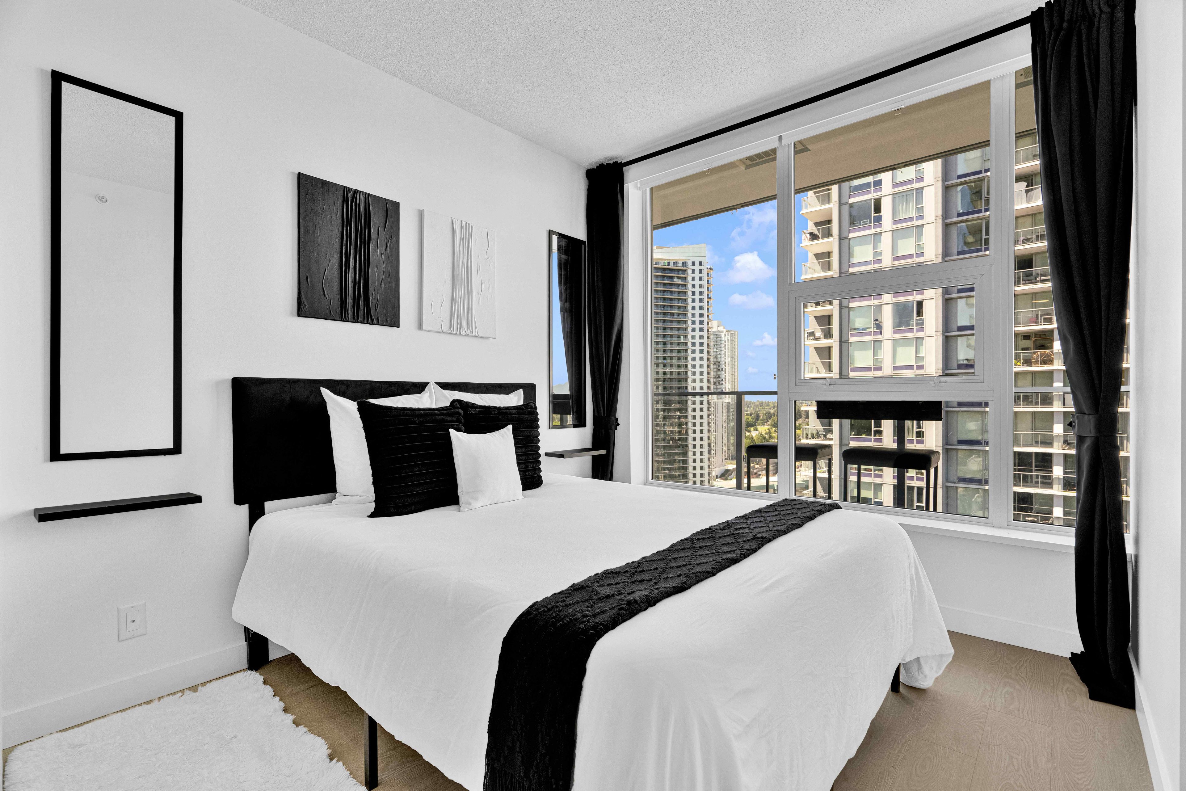 A modern bedroom with a large bed, black and white bedding, a view of buildings through a window, and artwork on the wall.