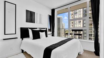 A modern bedroom with a large bed, black and white bedding, a view of buildings through a window, and artwork on the wall.