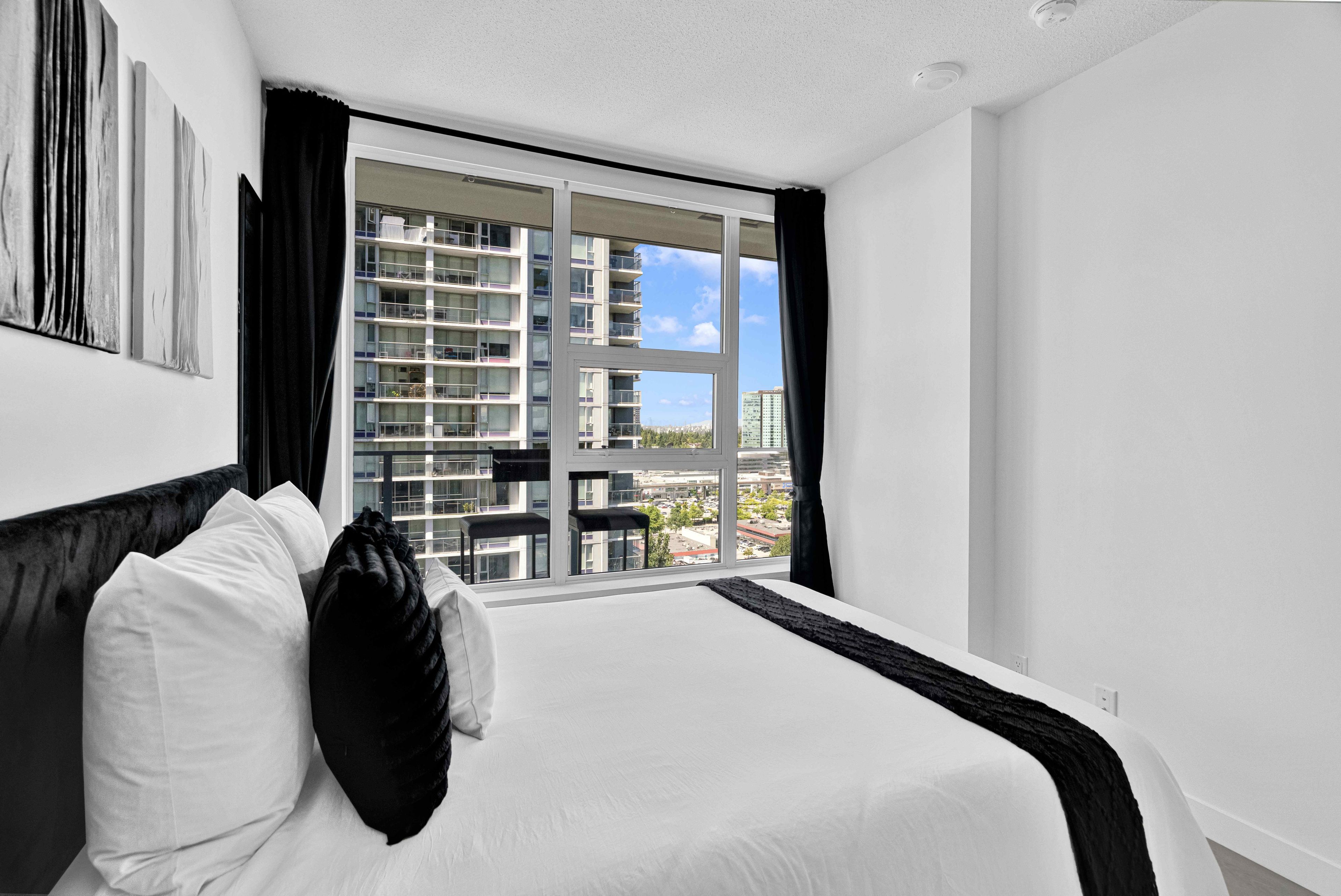 A hotel room with a large bed, a view of a high-rise building, and a balcony with outdoor furniture.