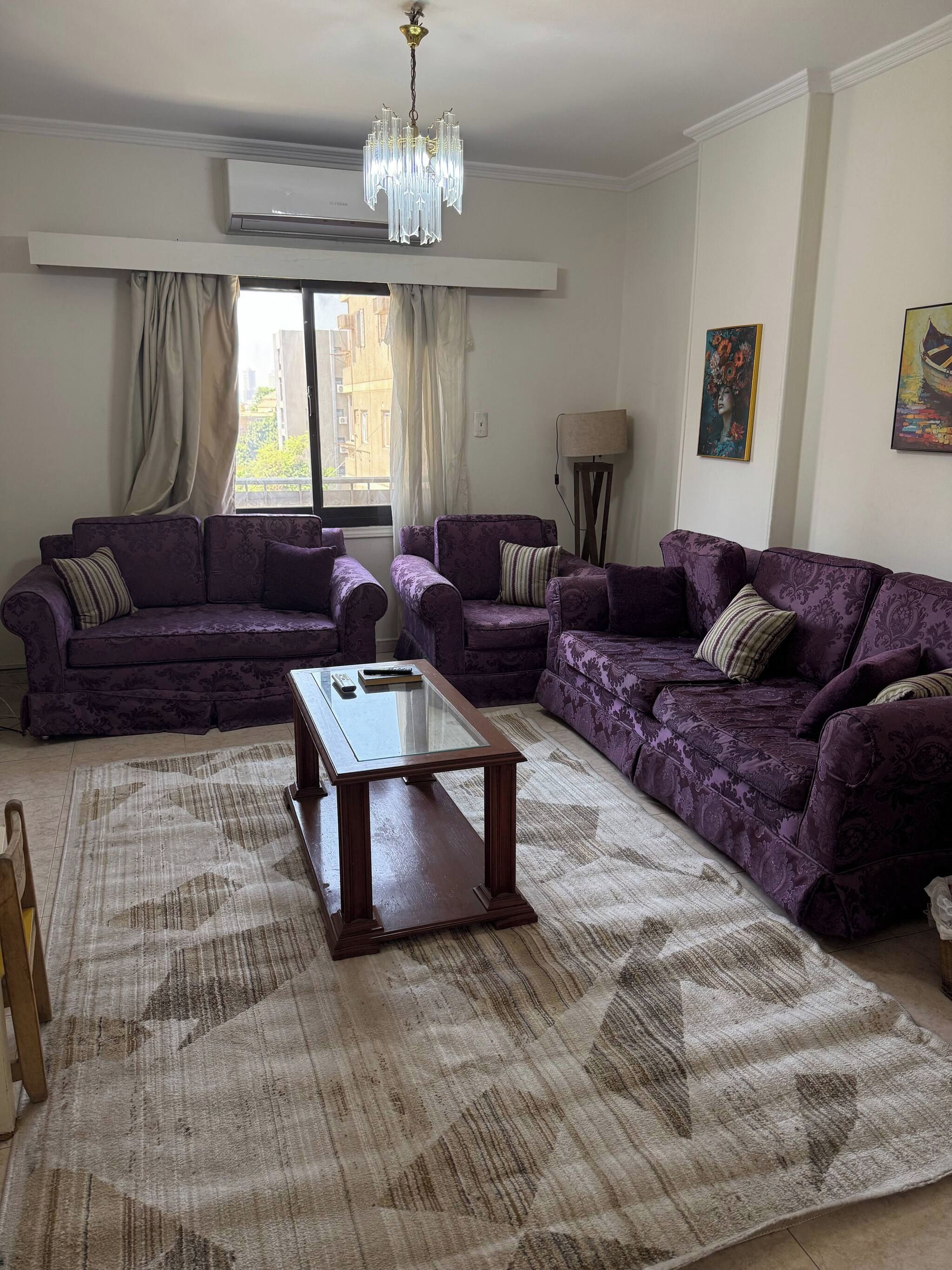 A living room with a large window, purple sofas, a glass-top coffee table, and a patterned rug.