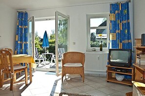 Ein Zimmer mit einem Esstisch, Stühlen, einer Korbschaukel, einem Fernseher auf einem Holzboden und einem Fenster mit Vorhängen.