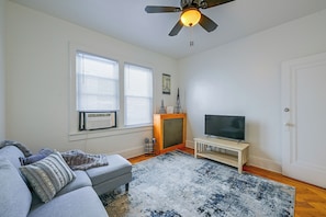 A living room with a grey sofa, a television on a wooden stand, a ceiling fan, and a window with blinds.