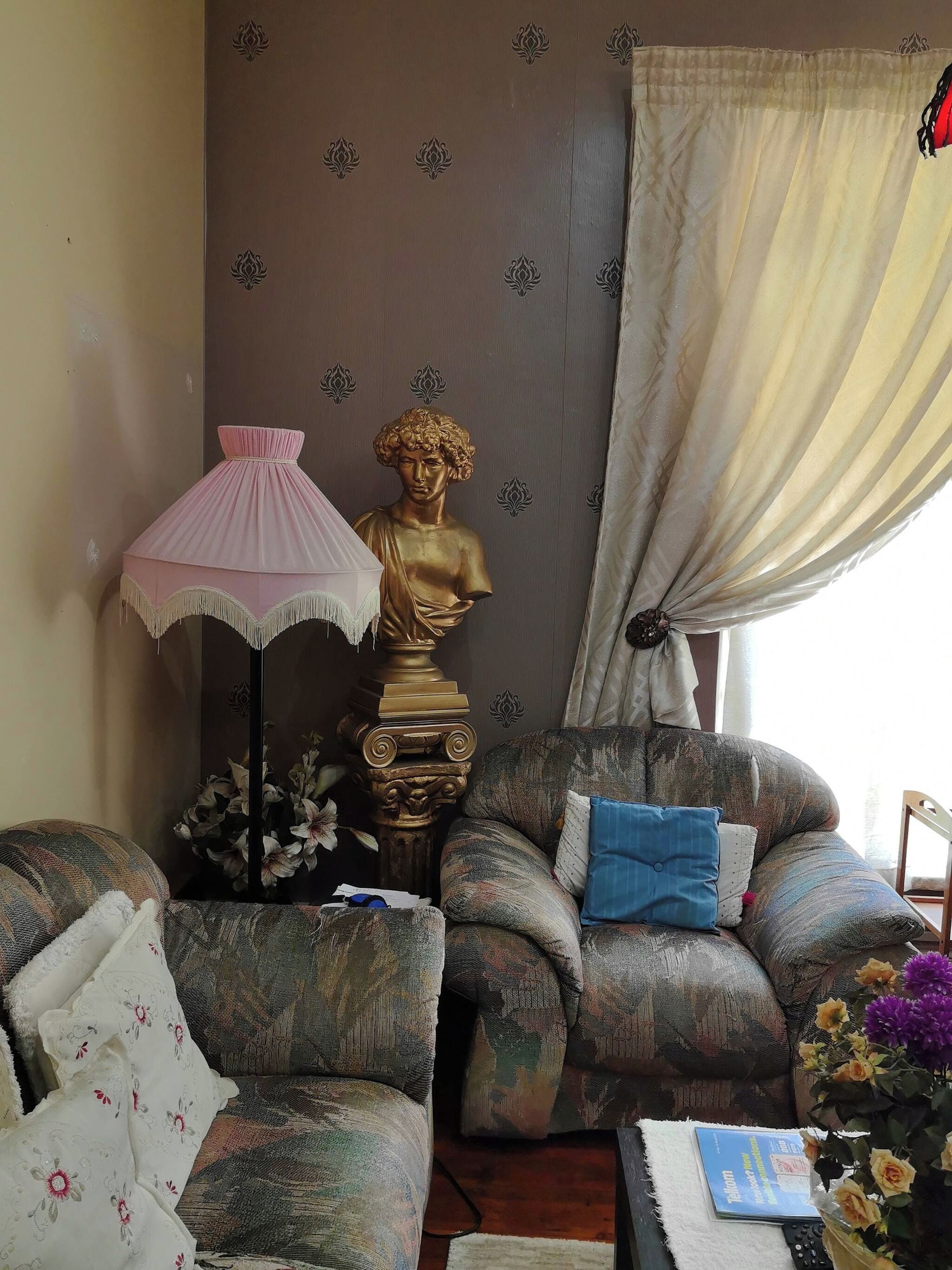Un salon avec un canapé, une lampe, une sculpture en buste et une fenêtre avec des rideaux.