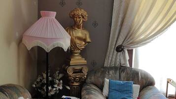 Un salon avec un canapé, une lampe, une sculpture en buste et une fenêtre avec des rideaux.