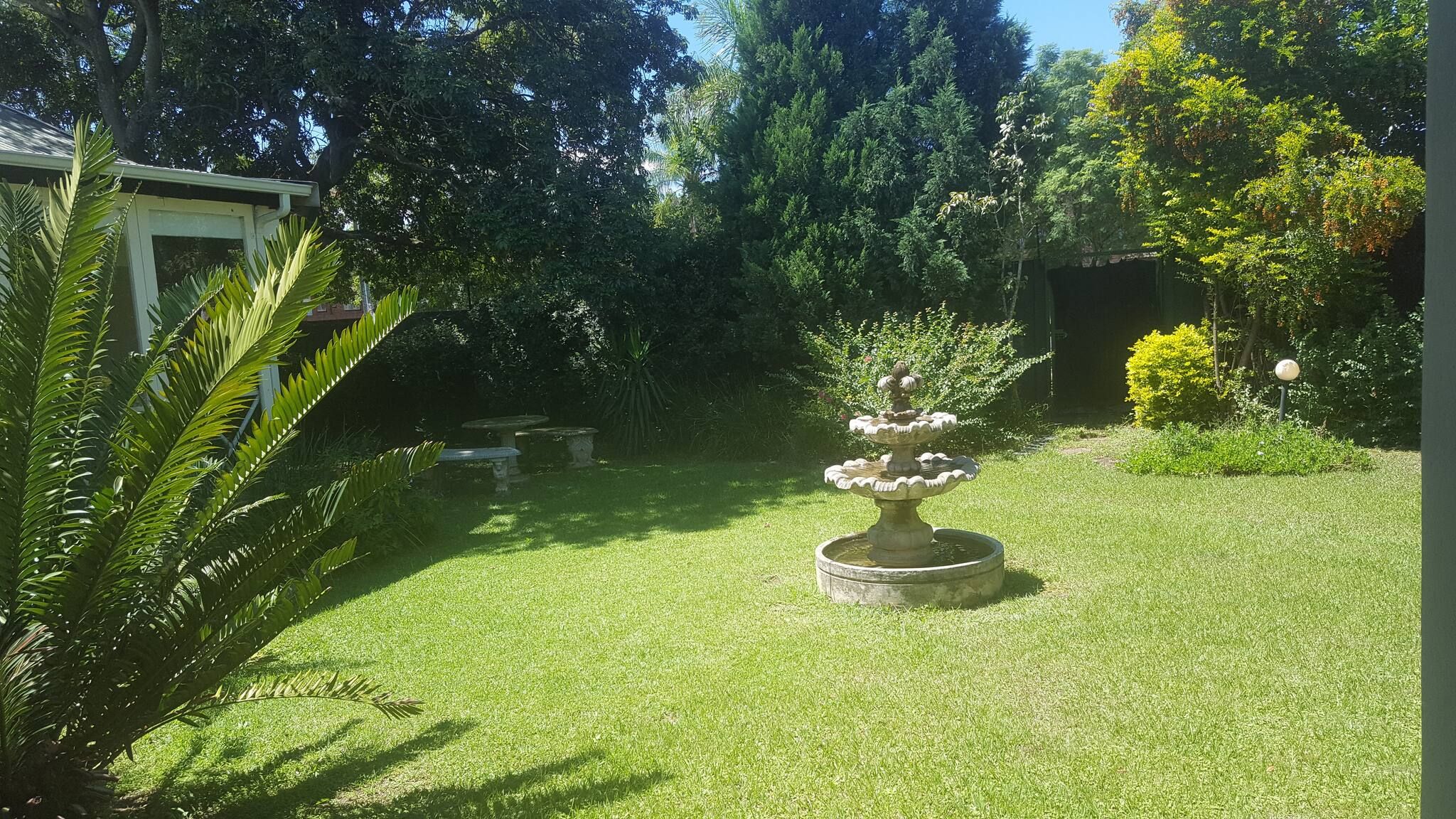 Un jardin bien entretenu, avec une fontaine centrale, entouré d’une végétation luxuriante et de diverses espèces d’arbres.