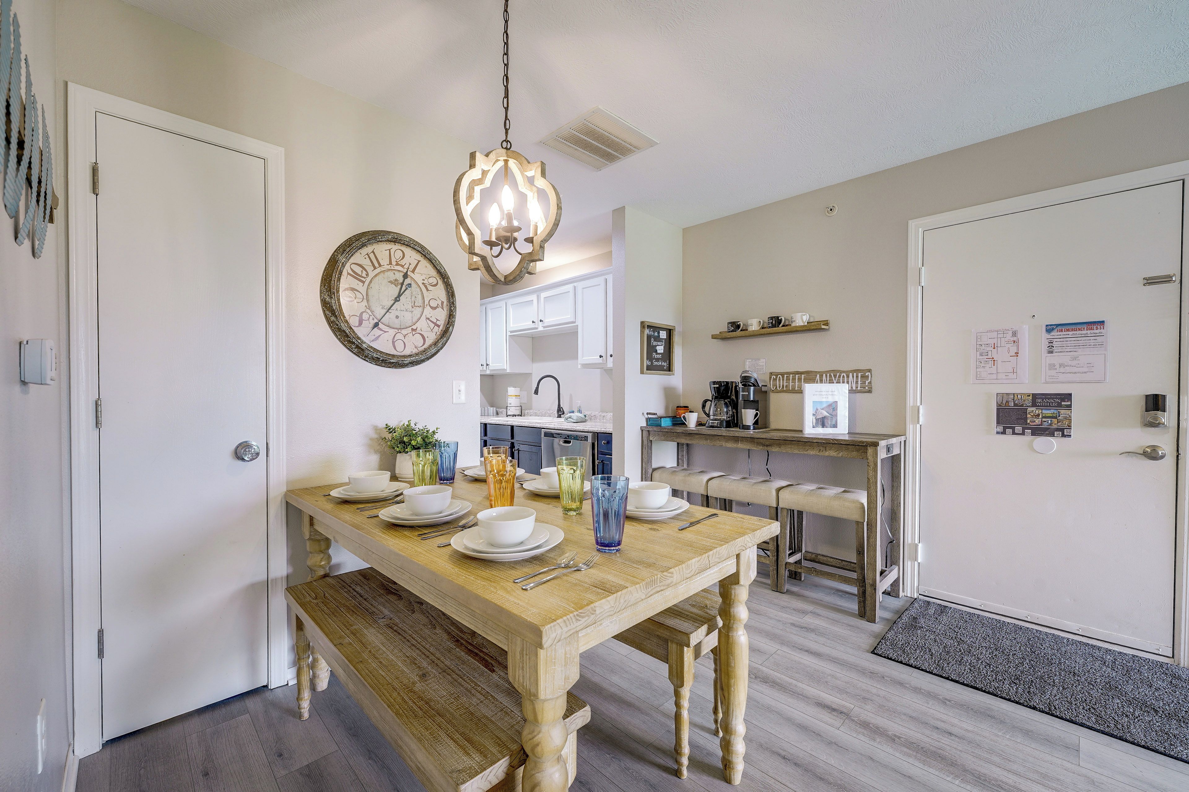 Un coin repas avec une table en bois dressée pour quatre personnes, une horloge au mur et une cuisine en arrière-plan.