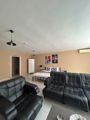 A living room with two black leather sofas, a bed with a white headboard, a white wardrobe, and a ceiling fan.