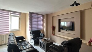 Ein Wohnzimmer mit einer Sofagarnitur, einem Couchtisch, einem an der Wand befestigten Fernseher und einem Deckenventilator.