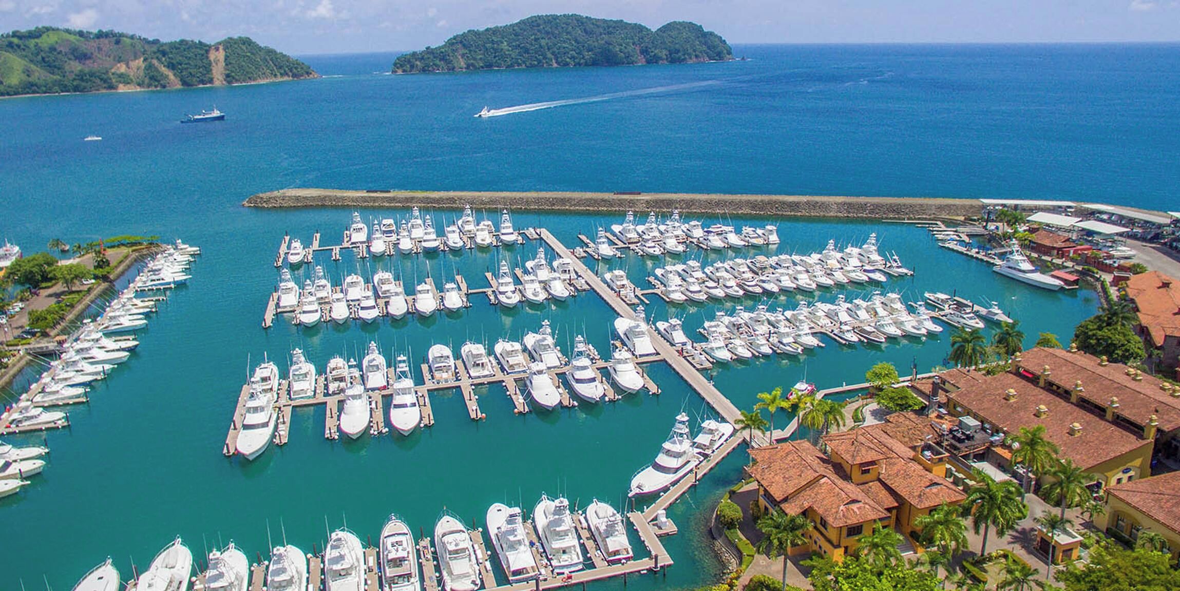 A marina with numerous boats docked in a structured layout, surrounded by a coastal landscape with hills in the background.