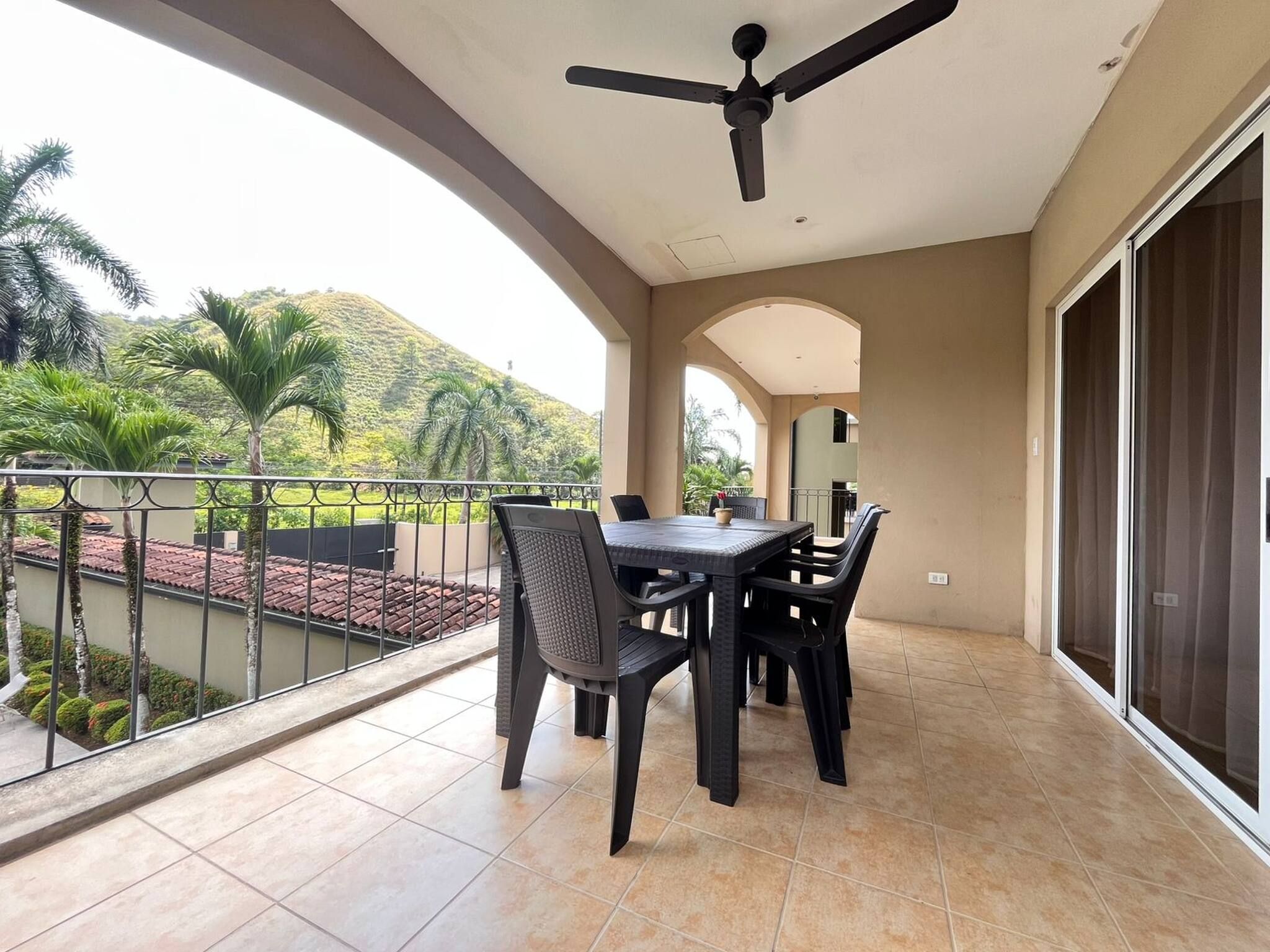 Un balcon avec une table et des chaises, un ventilateur de plafond et une vue sur une colline.
