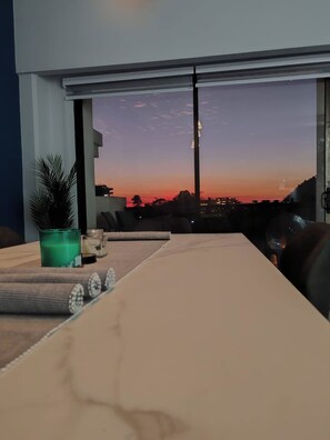 A room with a large window, a table with decorative items, and a view of a sunset and buildings. - Modern 3BR Condo in JacoBay - Walk to the beach 2blocks (Jaco)