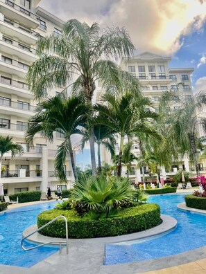 A hotel with a swimming pool surrounded by palm trees and a multi-story building with balconies. - Modern 3BR Condo in JacoBay - Walk to the beach 2blocks (Jaco)