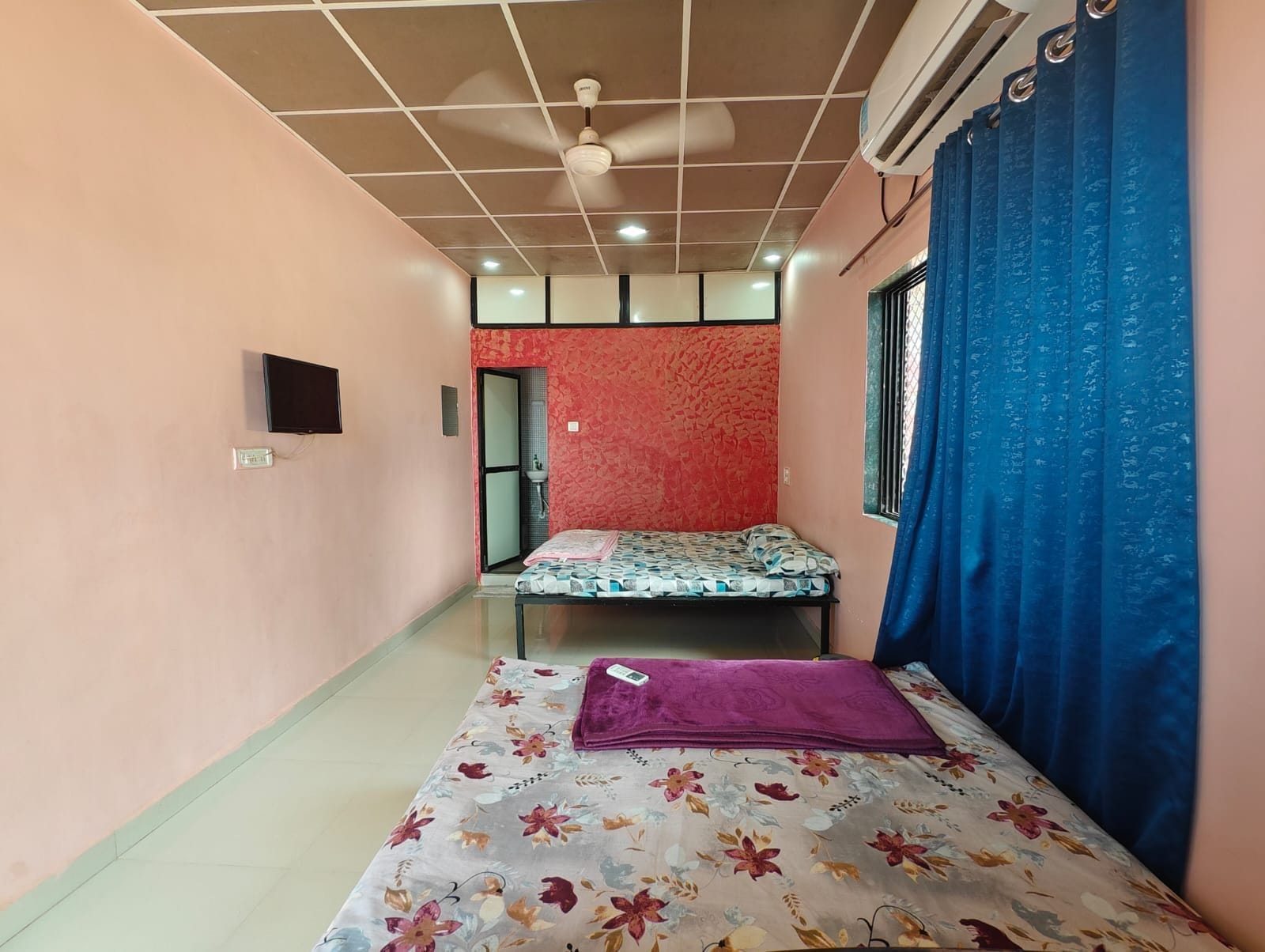Une petite pièce avec un lit, un ventilateur de plafond, une fenêtre avec des rideaux bleus et un téléviseur fixé au mur.