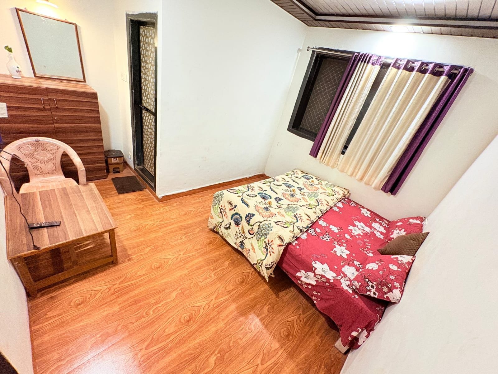 Una habitación pequeña con piso de madera, una cama con ropa de cama floral, una silla, un escritorio con un espejo y una ventana con cortinas.