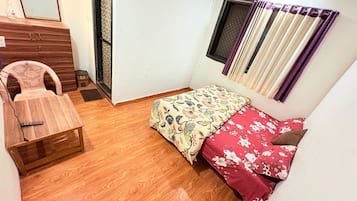 Una habitación pequeña con piso de madera, una cama con ropa de cama floral, una silla, un escritorio con un espejo y una ventana con cortinas.