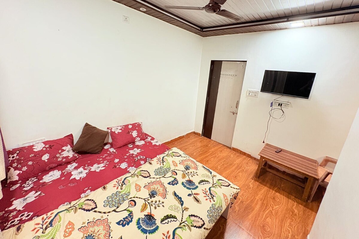 A bedroom with a bed, a wooden floor, a ceiling fan, a TV mounted on the wall, and a door.