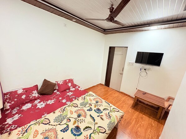 A bedroom with a bed, a wooden floor, a ceiling fan, a TV mounted on the wall, and a door.