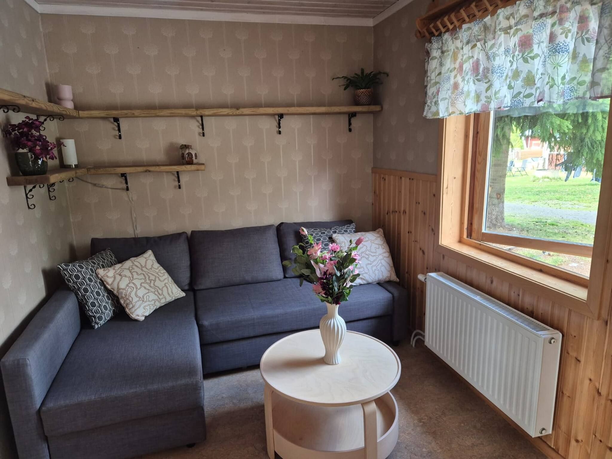 Ett mysigt rum med en grå hörnsoffa, ett litet runt bord med en vas med blommor och ett fönster med utsikt över grönskan.