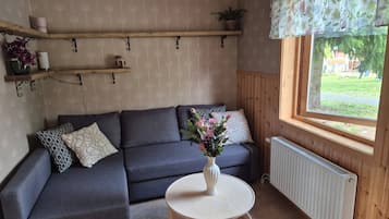 Ett mysigt rum med en grå hörnsoffa, ett litet runt bord med en vas med blommor och ett fönster med utsikt över grönskan.