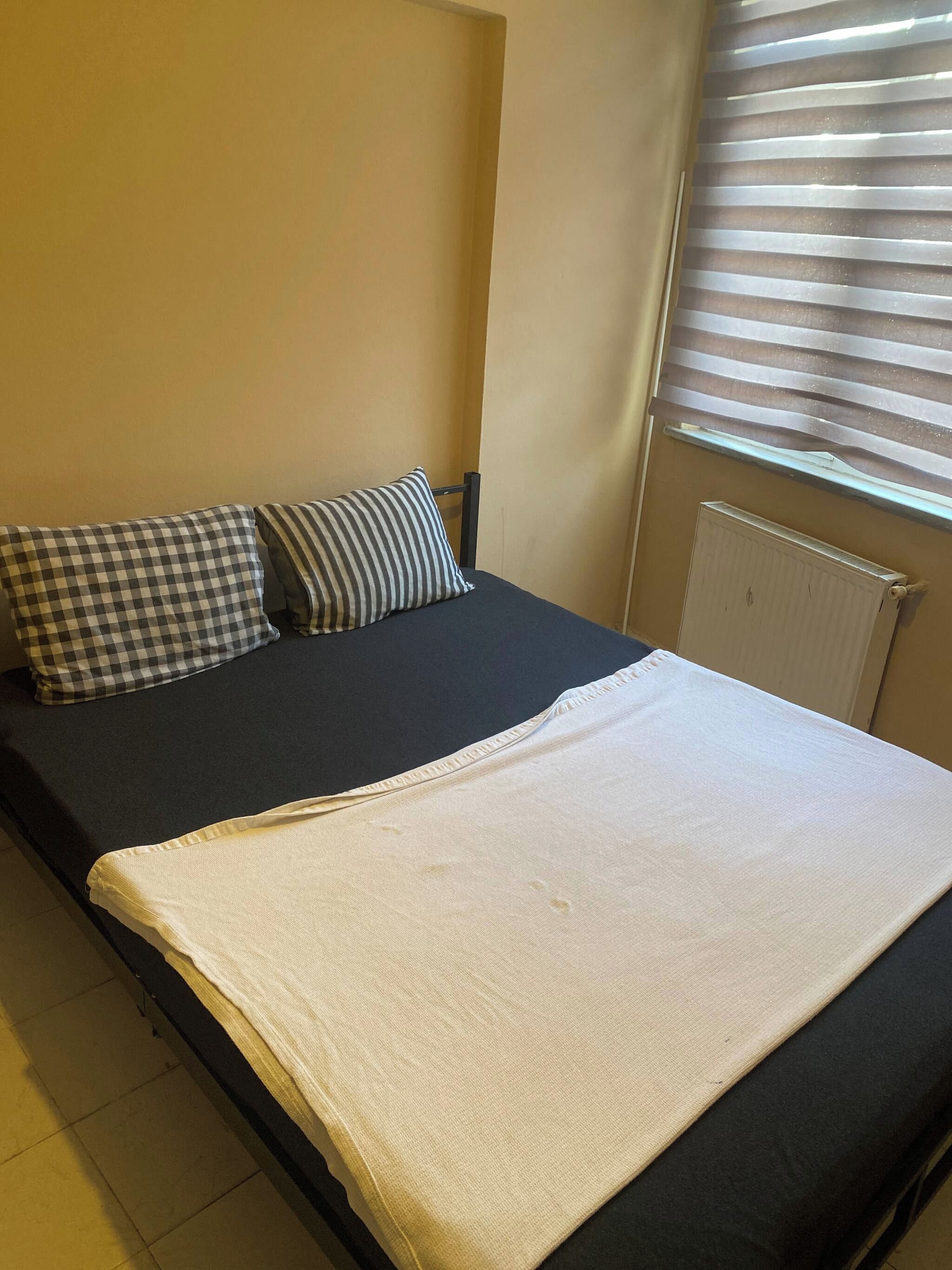 A single bed with a checkered pillow and a striped pillow, a white blanket, and a window with blinds.