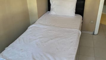 Una cama individual con colcha blanca y cabecera negra en una habitación con paredes color beige y suelo de baldosas.