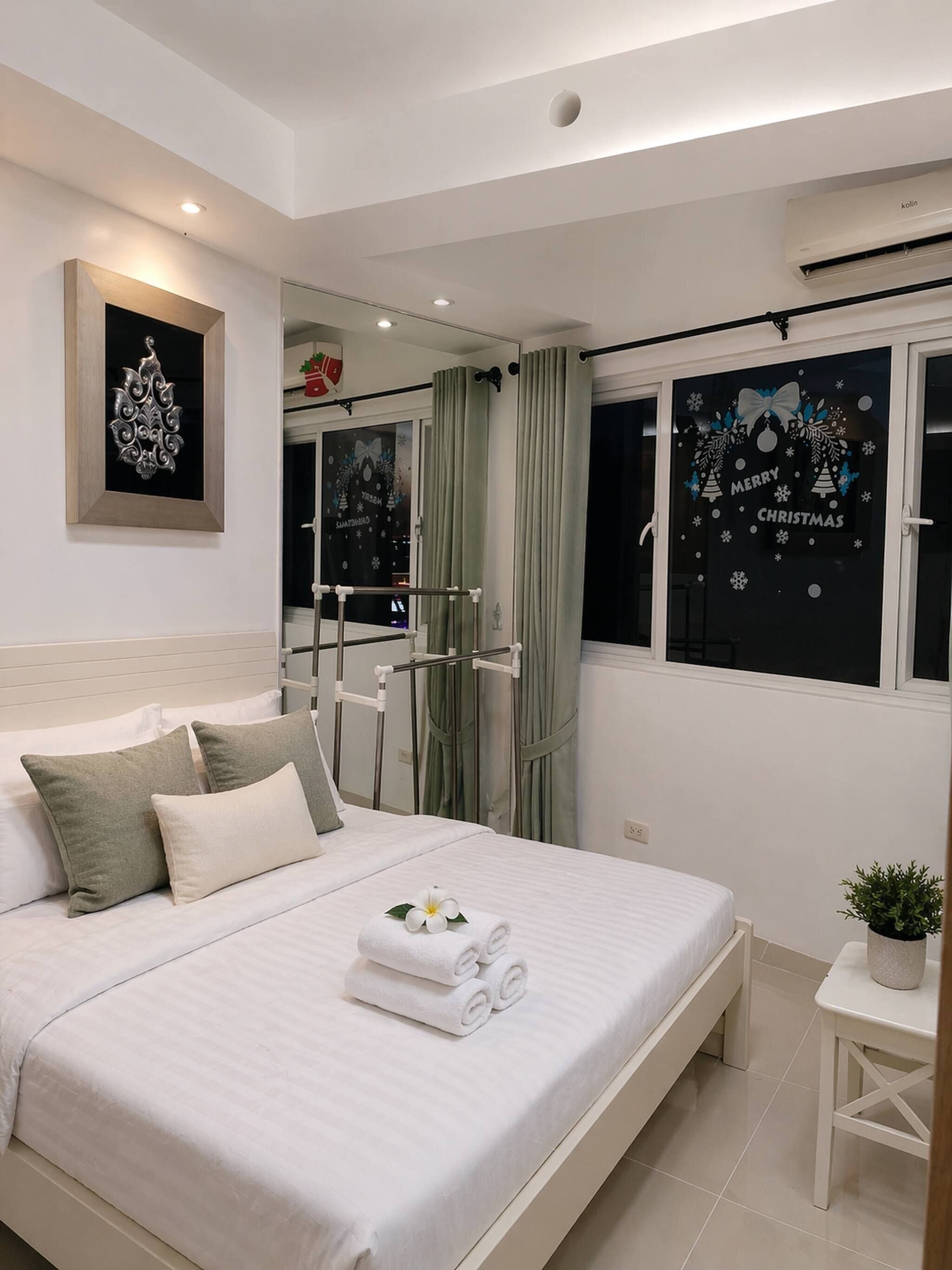 A hotel room with a bed, a small table with a plant, a mirror, and a window with a Christmas decoration.