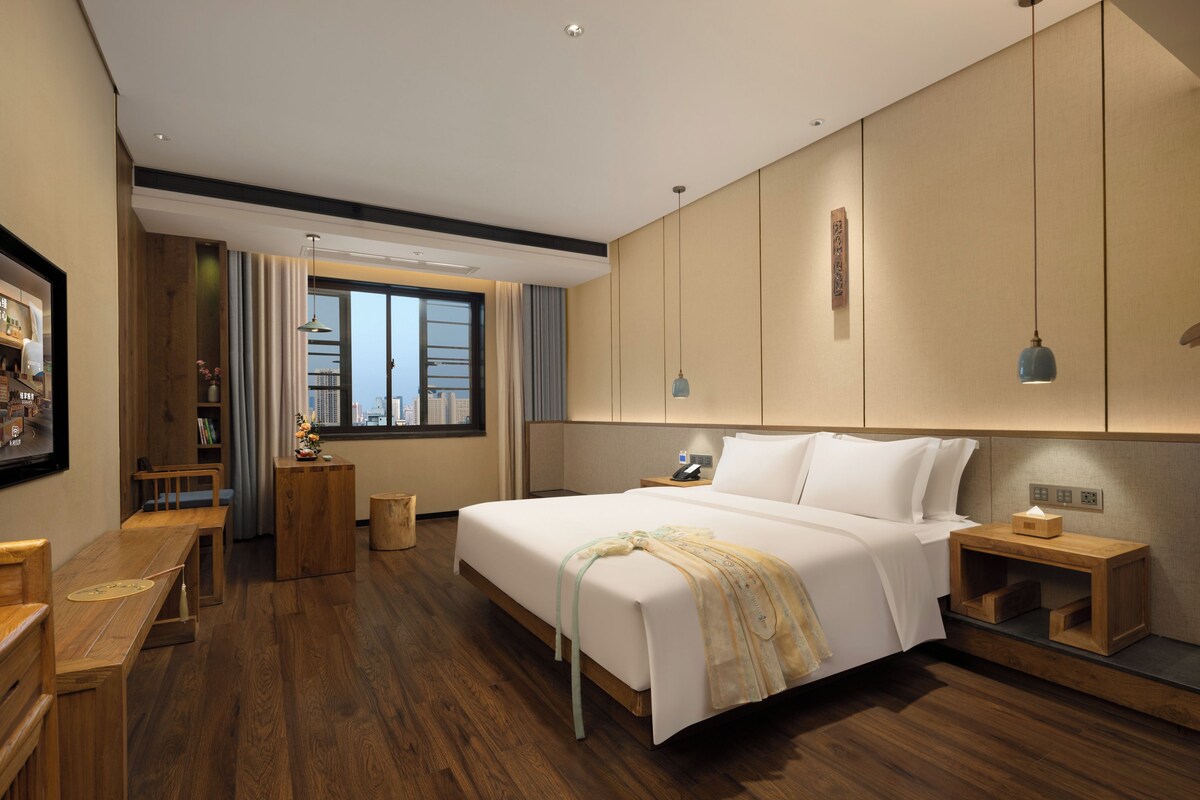 A modern hotel room with a large bed, wooden flooring, a desk, and a view of the city through the window.