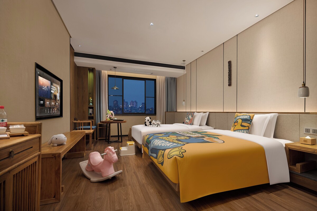 A hotel room with a large bed, a desk, a chair, a small table, and a window with a city view.