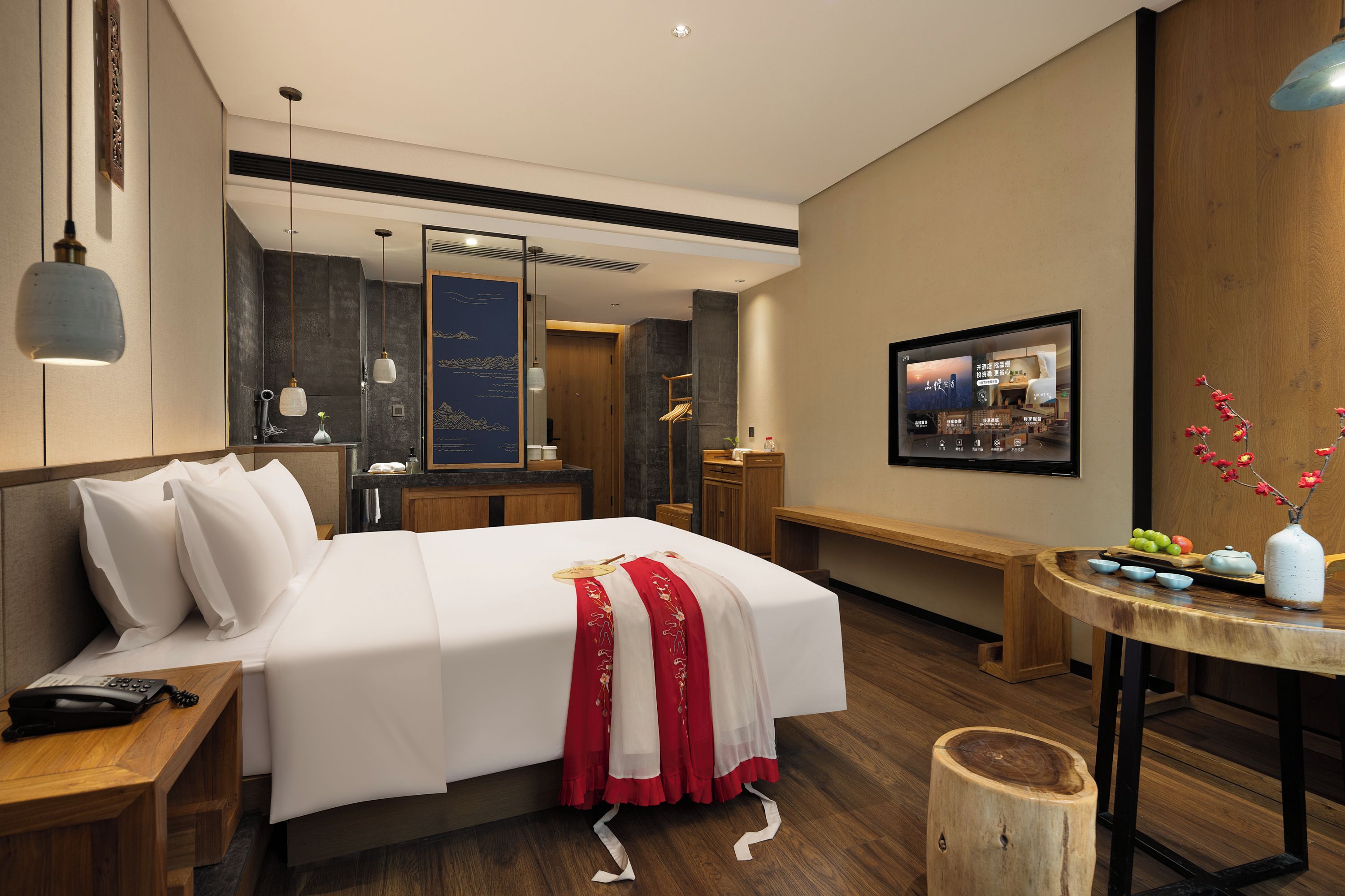 Quarto de hotel com uma cama grande, mesa de cabeceira, escrivaninha, cadeira, mesa redonda pequena com um vaso e televisão fixada na parede.