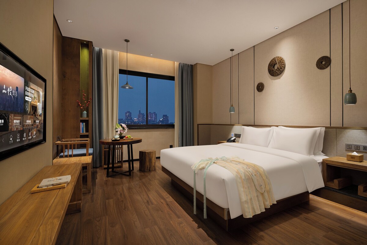 A hotel room with a large bed, a desk, a chair, a small table, and a window with a city view.