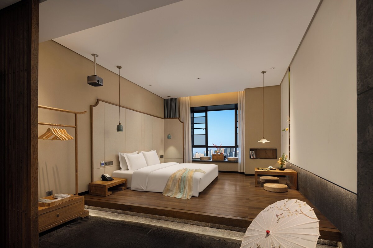 A modern hotel room with a large bed, wooden flooring, a desk, and a view of the city through a large window.