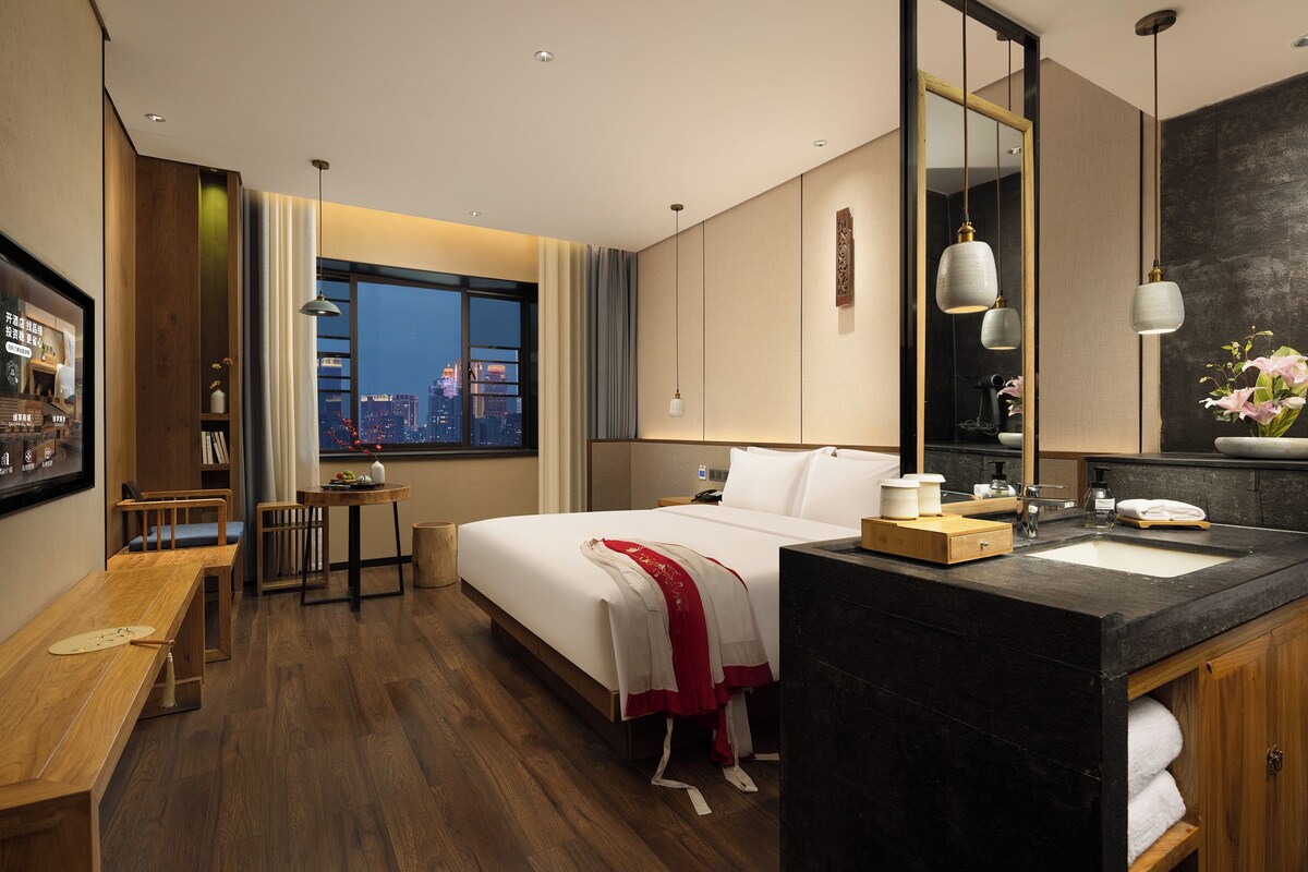 A hotel room with a bed, bedside table, a desk with a chair, a large window with a city view, a bathroom with a mirror, sink, and towel rack.