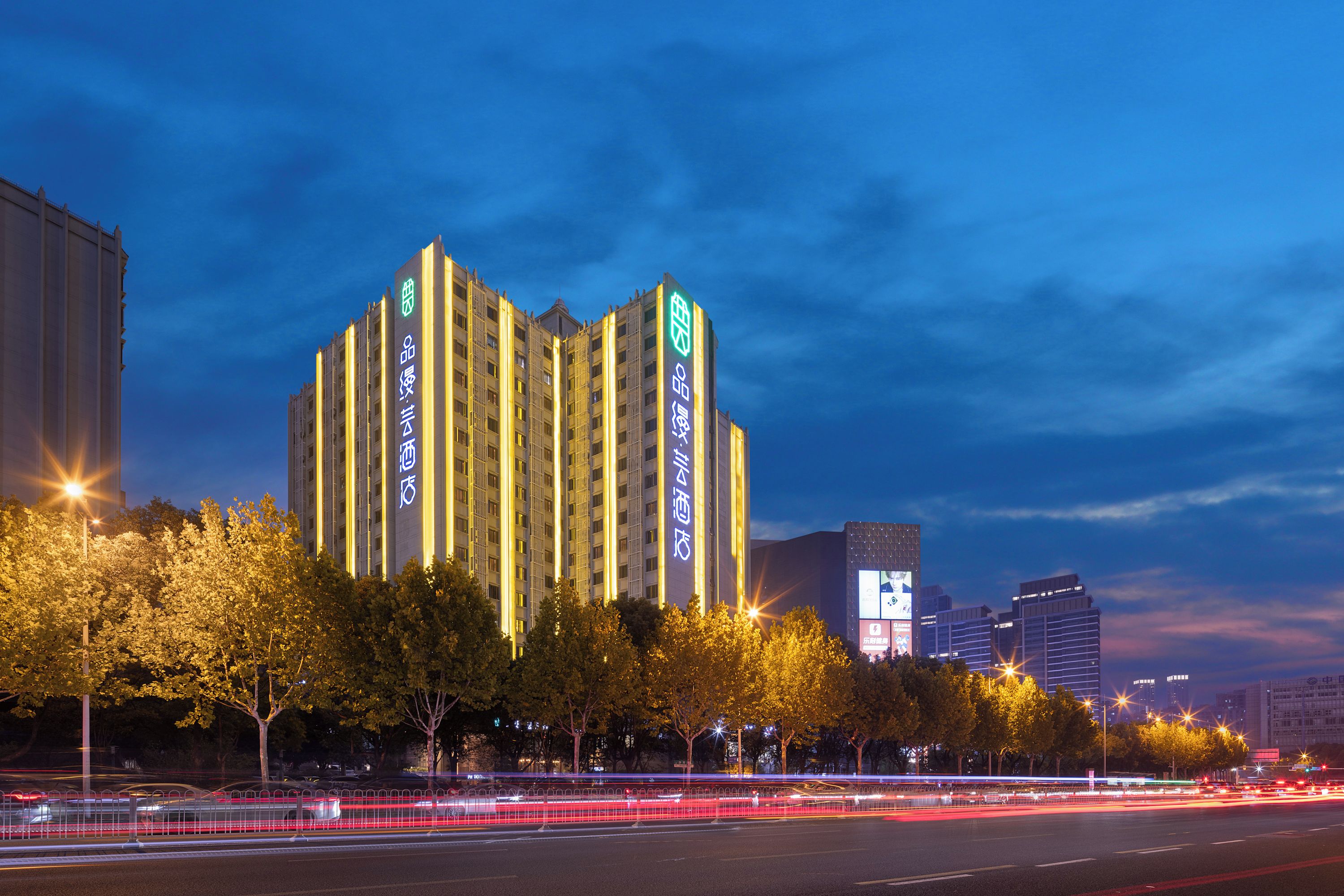Un hotel de varios pisos con señalización iluminada, rodeado de árboles y una calle concurrida con estelas de luz.