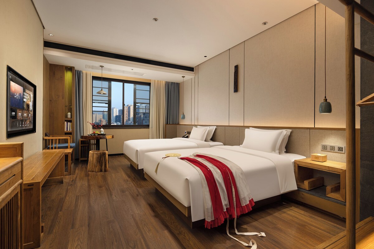A hotel room with two beds, a wooden desk, a small table with a vase, and a city view through the window.