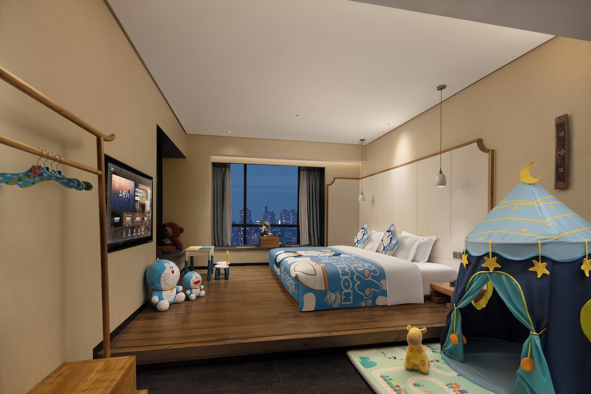 A hotel room with a city view, a bed, a desk, a chair, a small table, a teddy bear, and a tent-like play area.