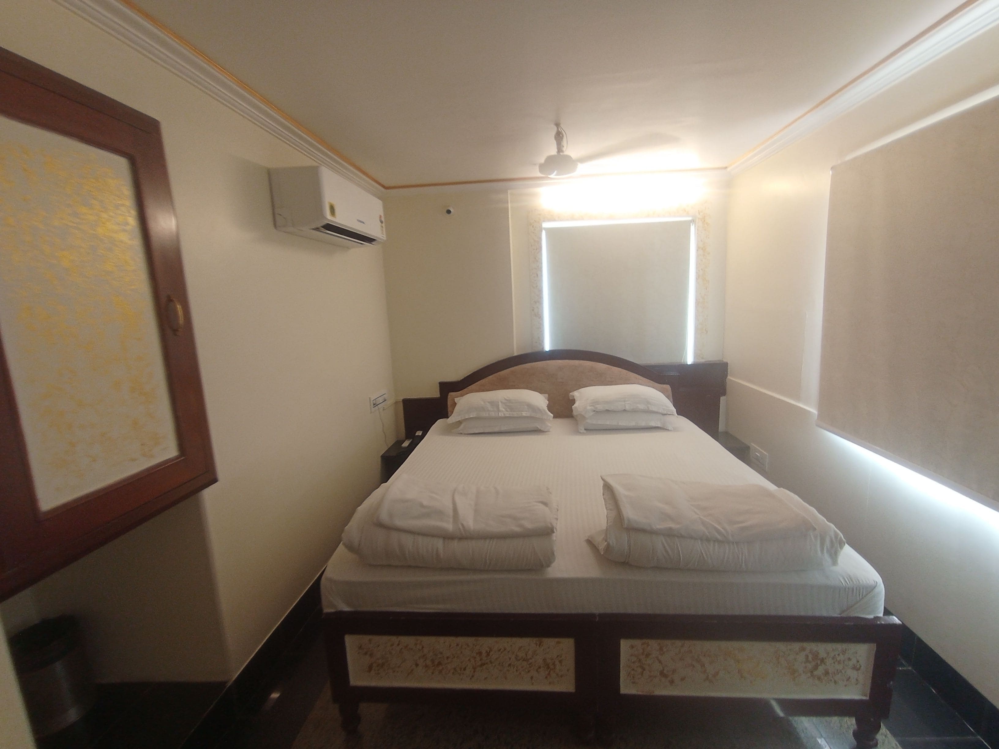 Een enkel bed met witte beddengoed in een kamer met plafondventilator, airconditioning en een raam met luxaflex.
