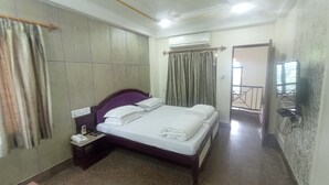 Quarto de hotel com cama, mesa de cabeceira, escrivaninha e uma cadeira. Há uma janela com cortinas, aparelho de ar condicionado e uma porta que dá para outro cômodo.