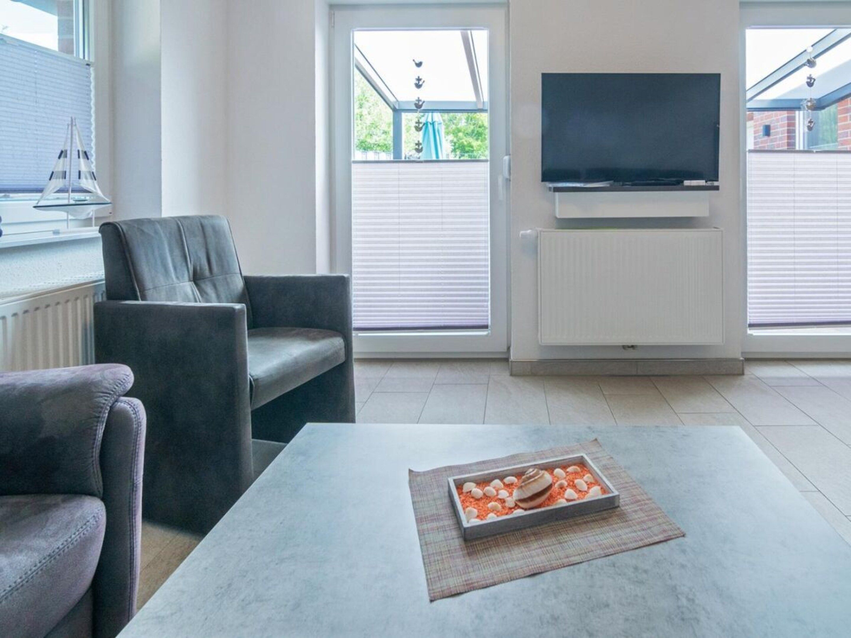 A modern living room with a sofa, a coffee table with a decorative item, a flat-screen TV mounted on the wall, and a window with blinds.