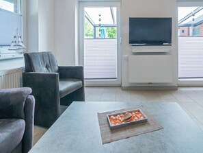 Ein modernes Wohnzimmer mit einer Couch, einem Couchtisch mit einer Dekoration, einem Flachbildfernseher an der Wand und einem Fenster mit Jalousien.