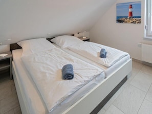 Ein Doppelbett mit weißen Bettlaken und zwei gerollten Handtüchern, ein Nachttisch mit Lampe, ein Fenster mit Blick auf einen Leuchtturm und ein Heizkörper an der Wand.