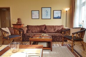 Ein Wohnzimmer mit einer Couch, zwei Sesseln, einem Couchtisch und einer Lampe. An der Wand hängen drei gerahmte Bilder.