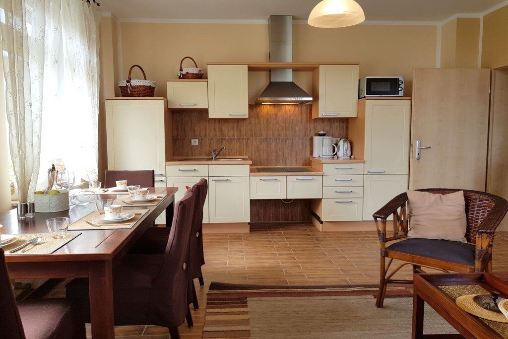 Une cuisine compacte avec des meubles blancs, un four à micro-ondes et un grille-pain. On y trouve une table à manger avec des chaises, ainsi qu’un fauteuil en osier.
