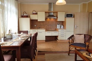 Eine kleine Küche mit weißen Schränken, einer Mikrowelle und einem Toaster. Es gibt einen Esstisch mit Stühlen und einen Korbstuhl.