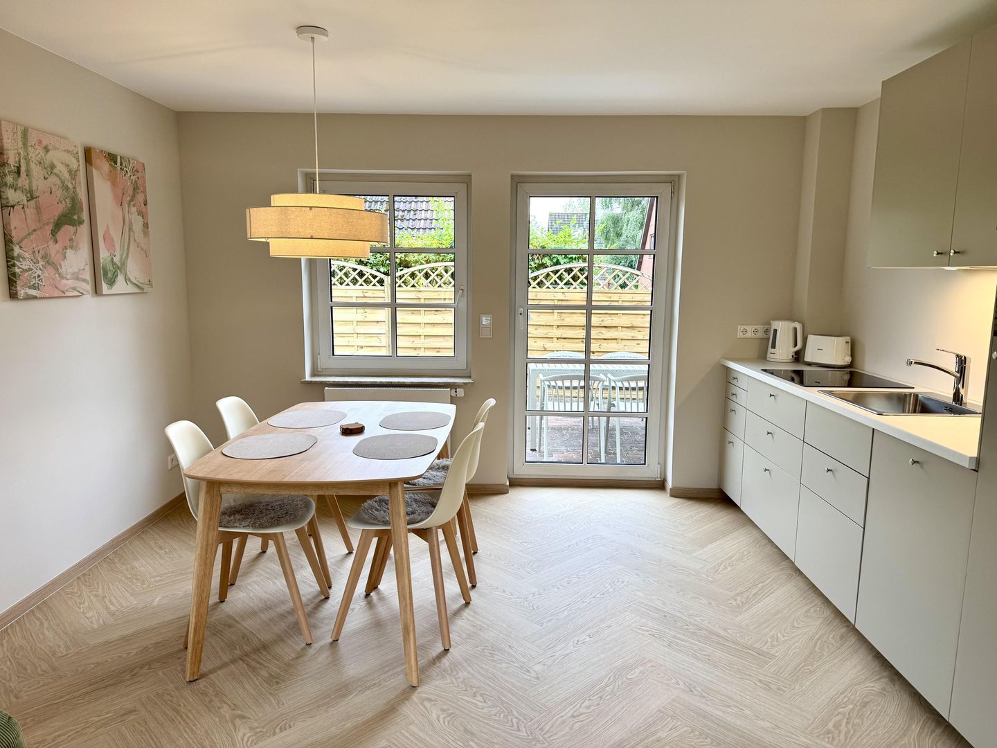 A modern kitchen with a dining area, featuring a table with chairs, a hanging light fixture, and a view through a glass door.